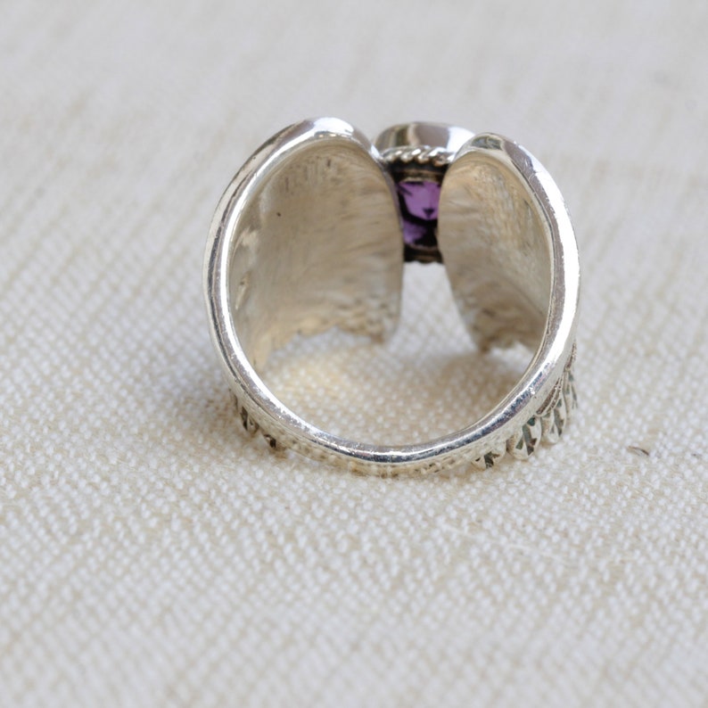May include: A silver ring with a purple gemstone set in the center. The ring has a detailed design on the band.