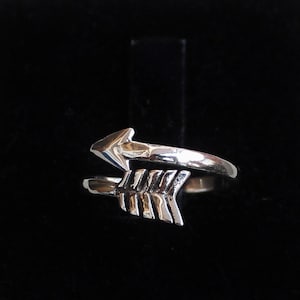 Puede incluir: Un anillo de plata con un diseño de flecha retorcida. La flecha apunta hacia la izquierda y tiene un diseño de plumas detallado.