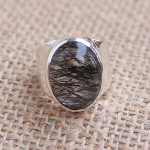 May include: A silver ring with an oval black and clear gemstone. The gemstone has a distinctive pattern of black inclusions, set in a polished silver band. The ring is displayed on a textured, neutral-toned surface.