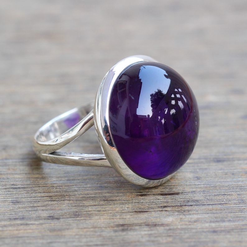 May include: A silver ring with a large, oval-shaped amethyst gemstone. The gemstone is a deep purple color and has a high polish.
