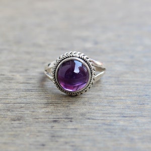 May include: A silver ring featuring a large, round, deep purple gemstone. The gemstone is set in a decorative bezel with a braided rope-like design. The ring has a split shank design.