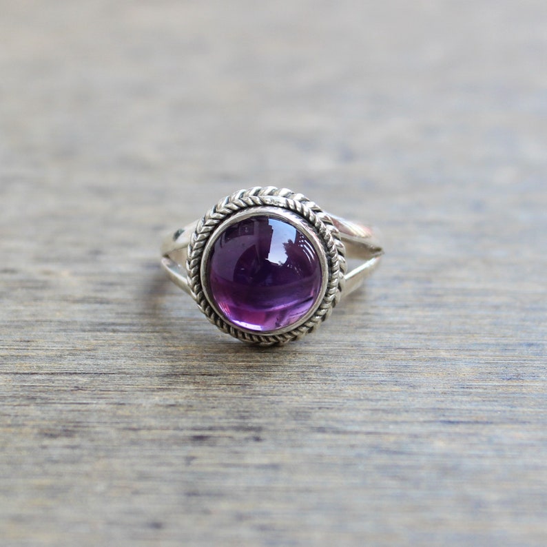 May include: A silver ring featuring a large, round, deep purple gemstone. The gemstone is set in a decorative bezel with a braided rope-like design. The ring has a split shank design.