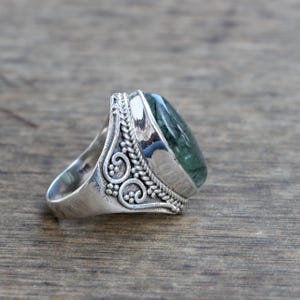 K&ouml;nnte beinhalten: Ein silberner Ring mit einem ovalen, gr&uuml;nen Edelstein, der in eine detaillierte Silberband gefasst ist. Die Band hat ein dekoratives Design mit Spiralen und Knoten.