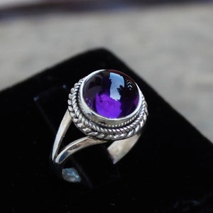 May include: A silver ring with a large, round, deep purple gemstone. The gemstone is set in a bezel with a twisted rope design around it. The ring has a split shank design.
