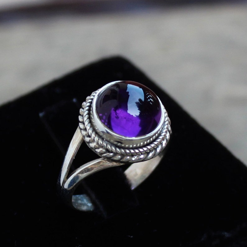 May include: A silver ring with a large, round, deep purple gemstone. The gemstone is set in a bezel with a twisted rope design around it. The ring has a split shank design.