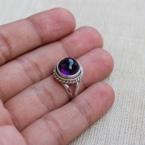 May include: A sterling silver ring featuring a large, round, deep purple amethyst gemstone. The gemstone is set in a decorative bezel with a twisted rope design. The ring's band splits into two strands as it approaches the gemstone.