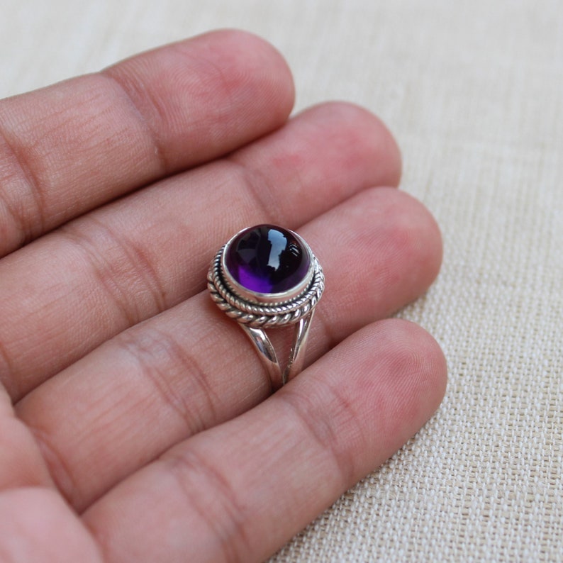 May include: A sterling silver ring featuring a large, round, deep purple amethyst gemstone. The gemstone is set in a decorative bezel with a twisted rope design. The ring's band splits into two strands as it approaches the gemstone.