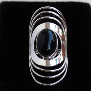 May include: A silver ring with a black oval gemstone set in a circular design. The ring has multiple concentric circles.