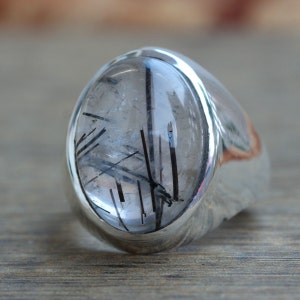 May include: A silver ring featuring a clear quartz stone with black tourmaline inclusions. The stone is oval shaped and set in a silver band.