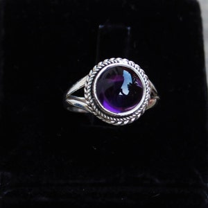 May include: A silver ring featuring a large, round, deep purple amethyst gemstone. The gemstone is set in a decorative bezel with a twisted rope design. The ring has a split shank design.