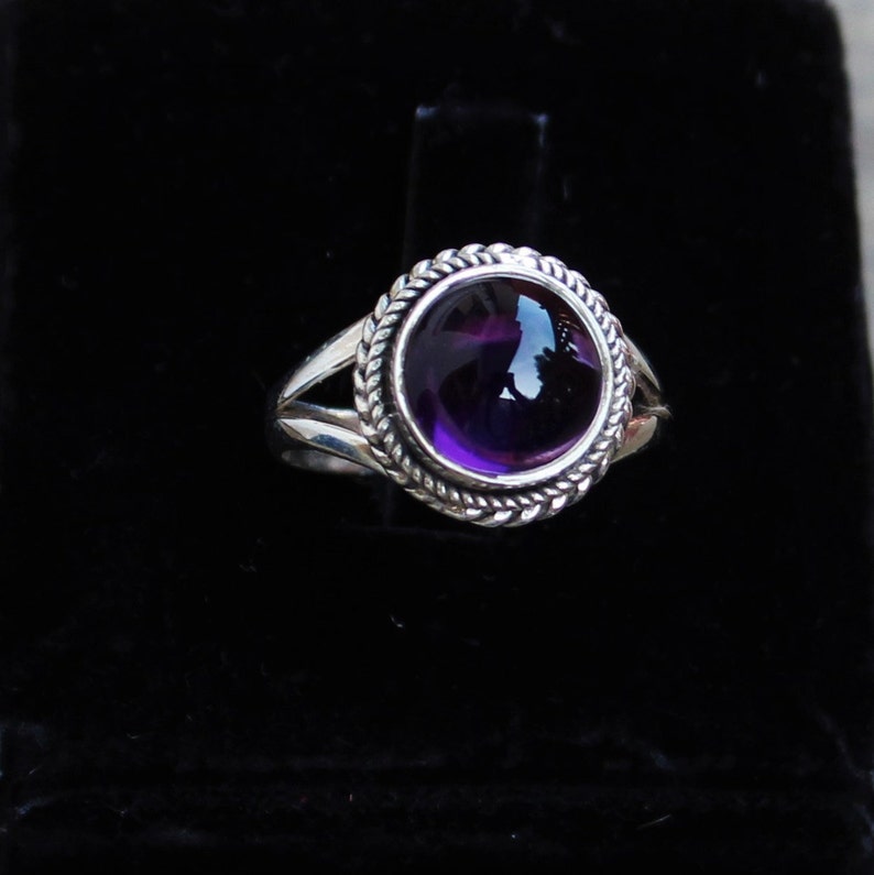 May include: A silver ring featuring a large, round, deep purple amethyst gemstone. The gemstone is set in a decorative bezel with a twisted rope design. The ring has a split shank design.