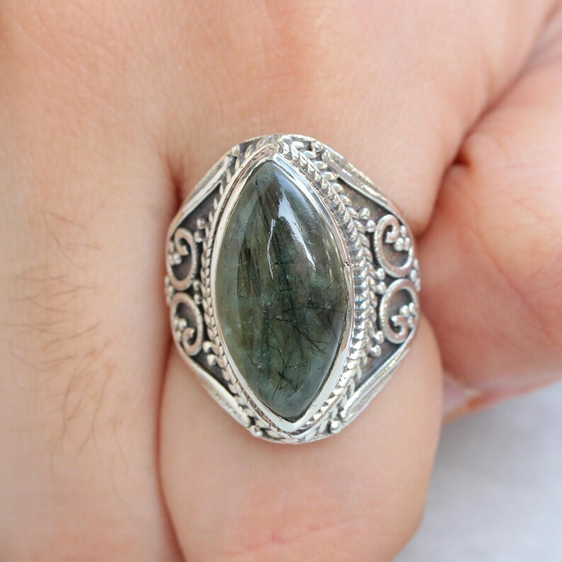 K&ouml;nnte beinhalten: Ein silberner Ring mit einem verzierten Design zeigt einen gro&szlig;en, dunkelgr&uuml;nen Edelstein mit einem marmorierten Muster.