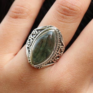 K&ouml;nnte beinhalten: Ein silberner Ring mit einem verzierten Design, das einen gr&uuml;nen Edelstein in der Mitte zeigt.