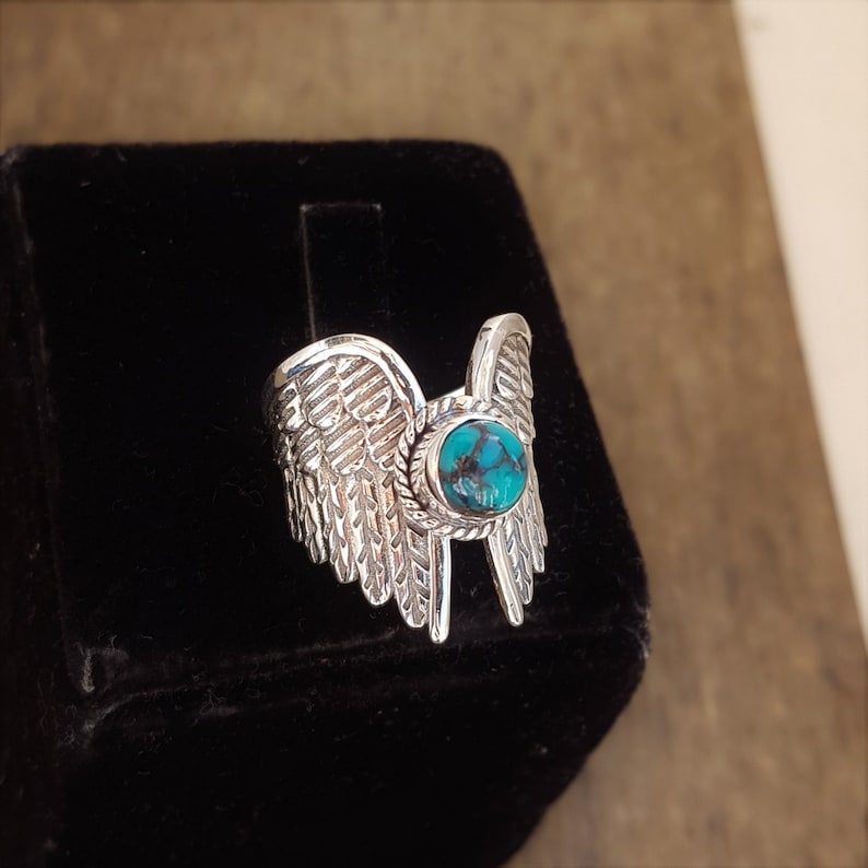 Op de afbeelding: Een zilveren ring met een turquoise steen in het midden, omringd door twee vleugels.