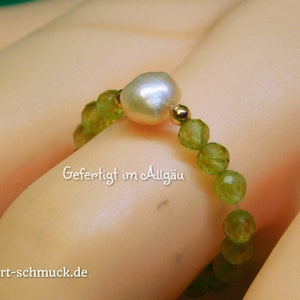 Könnte beinhalten: Ein Ring aus grünen Edelsteinperlen und einer weißen Perle. Der Ring befindet sich an einer Hand und der Text "Gefertigt im Allgäu" und "herzt-schmuck.de" ist sichtbar.