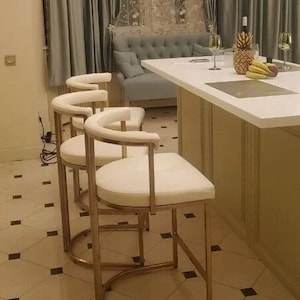 Pode incluir: Três banquetas de bar de metal dourado com assentos acolchoados brancos. As banquetas estão em uma sala com uma ilha de cozinha branca e um sofá azul claro.