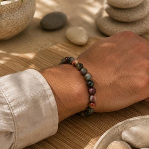Könnte beinhalten: Ein elastisches Armband bestehend aus kleinen, runden, polierten Edelsteinperlen in verschiedenen Erdtönen, darunter Schattierungen von tiefem Grün, gedämpftem Rot, Braun und Grau, getragen am Handgelenk mit einer glatten, glänzenden Oberfläche.