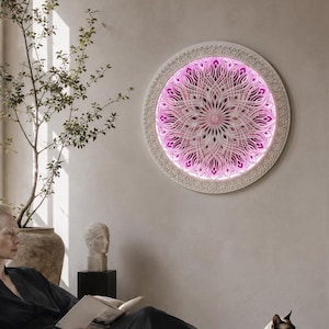 May include: A circular wall-mounted light fixture featuring an intricate, layered mandala pattern in white, illuminated from behind with a vibrant pink glow. The design consists of symmetrical, radiating geometric cutouts that create depth.