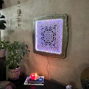 Pu&ograve; includere: Un'opera d'arte murale quadrata e multistrato con un intricato design geometrico. L'opera &egrave; illuminata da una morbida luce viola, creando un effetto visivo accattivante. Il pezzo &egrave; montato su una parete strutturata dai toni neutri.