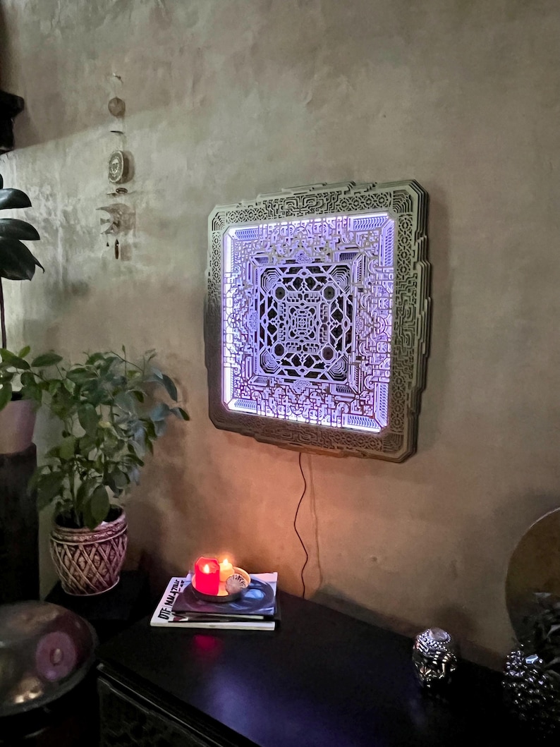 Pu&ograve; includere: Un'opera d'arte murale quadrata e multistrato con un intricato design geometrico. L'opera &egrave; illuminata da una morbida luce viola, creando un effetto visivo accattivante. Il pezzo &egrave; montato su una parete strutturata dai toni neutri.