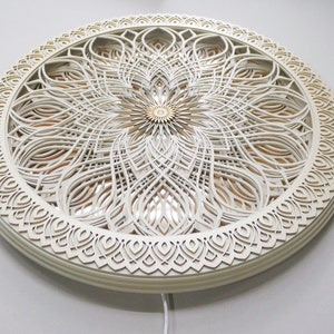 May include: A circular wall art piece featuring multiple layers of white laser-cut wood arranged in a symmetrical floral pattern. The center has a small gold-toned accent, and the design includes intricate cutouts that reveal a warm light source.