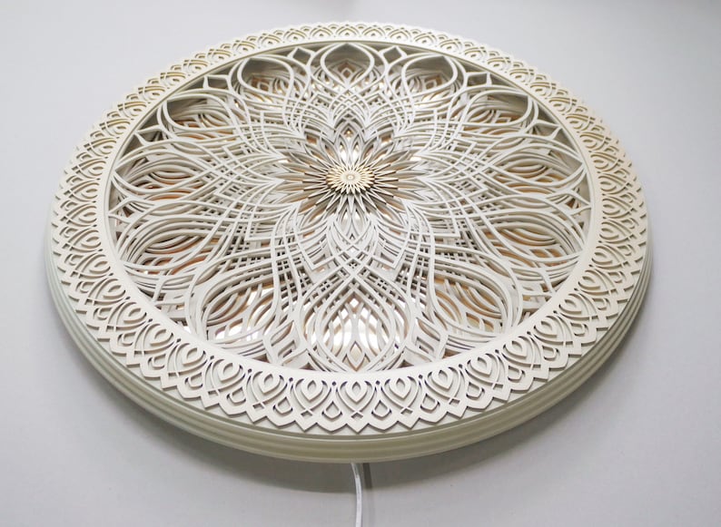 May include: A circular wall art piece featuring multiple layers of white laser-cut wood arranged in a symmetrical floral pattern. The center has a small gold-toned accent, and the design includes intricate cutouts that reveal a warm light source.