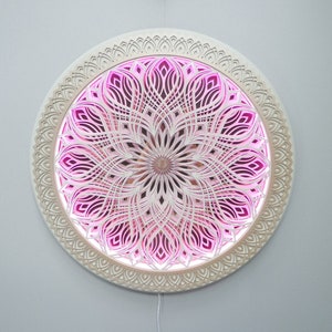May include: A circular wall-mounted art piece featuring intricate, layered white laser-cut patterns arranged in a symmetrical mandala design. The center and inner sections are illuminated with a vibrant pink light, creating depth and contrast.