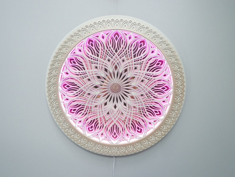May include: A circular wall-mounted art piece featuring intricate, layered white laser-cut patterns arranged in a symmetrical mandala design. The center and inner sections are illuminated with a vibrant pink light, creating depth and contrast.