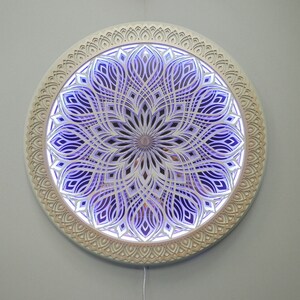 May include: A circular wall-mounted light fixture featuring intricate, layered, laser-cut wooden panels arranged in a symmetrical mandala pattern. The design displays a series of concentric, petal-like cutouts illuminated from behind by a vibrant purple glow.