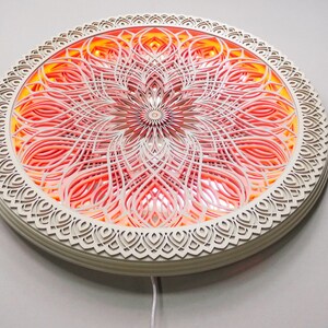 May include: A circular wall-mounted light fixture featuring intricate, layered laser-cut wood panels in a mandala pattern. The design displays concentric shapes with a warm, glowing orange and pink light emanating from behind the cutouts.