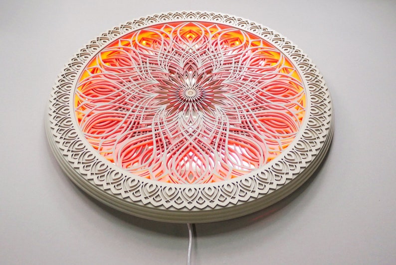 May include: A circular wall-mounted light fixture featuring intricate, layered laser-cut wood panels in a mandala pattern. The design displays concentric shapes with a warm, glowing orange and pink light emanating from behind the cutouts.