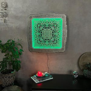 Pu&ograve; includere: Un'opera d'arte da parete quadrata dal design complesso con un interno verde luminoso. L'opera d'arte &egrave; montata su un muro grigio e il design presenta motivi geometrici. Una piccola candela e dei libri sono su una superficie nera sottostante.