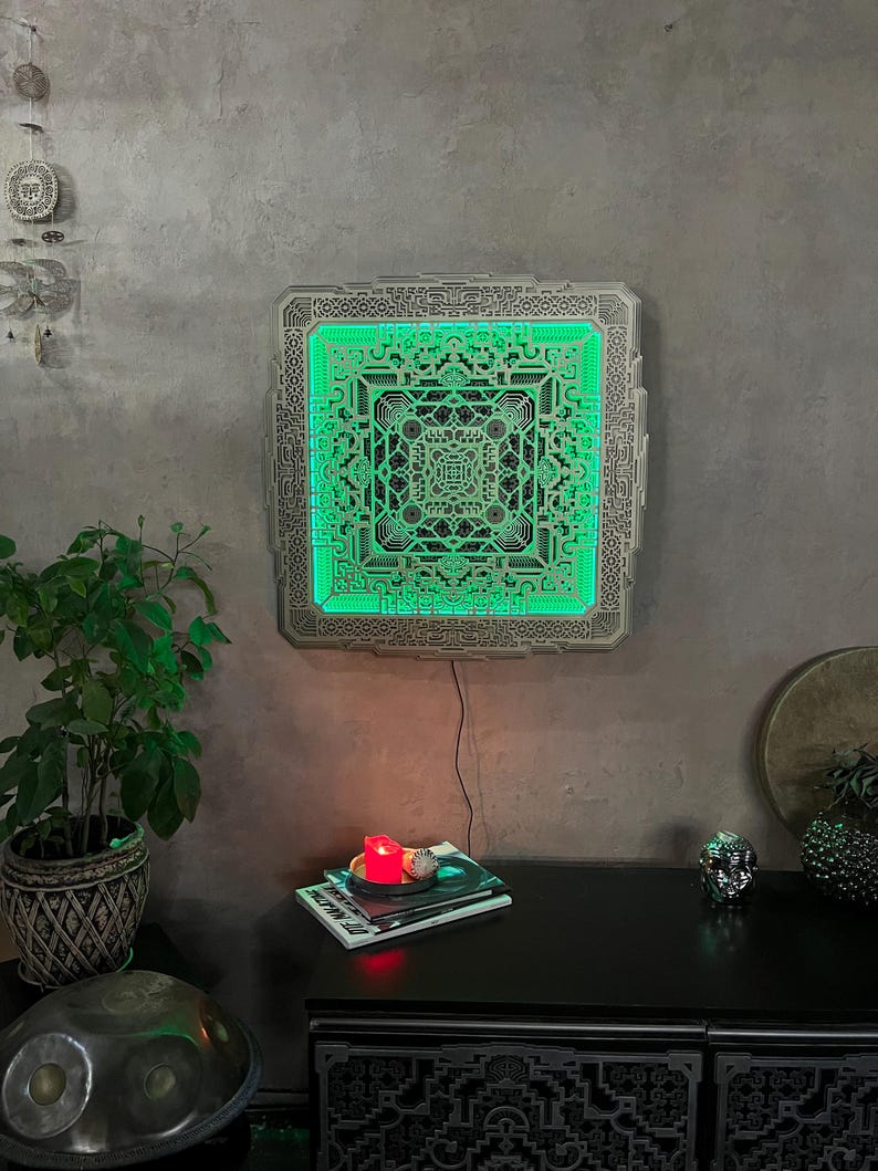 Pu&ograve; includere: Un'opera d'arte da parete quadrata dal design complesso con un interno verde luminoso. L'opera d'arte &egrave; montata su un muro grigio e il design presenta motivi geometrici. Una piccola candela e dei libri sono su una superficie nera sottostante.