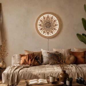 May include: A circular wall-mounted light fixture featuring an intricate, laser-cut mandala pattern in a light wood finish. The design is backlit, emitting a warm, soft glow through the detailed geometric cutouts, creating a decorative focal point.