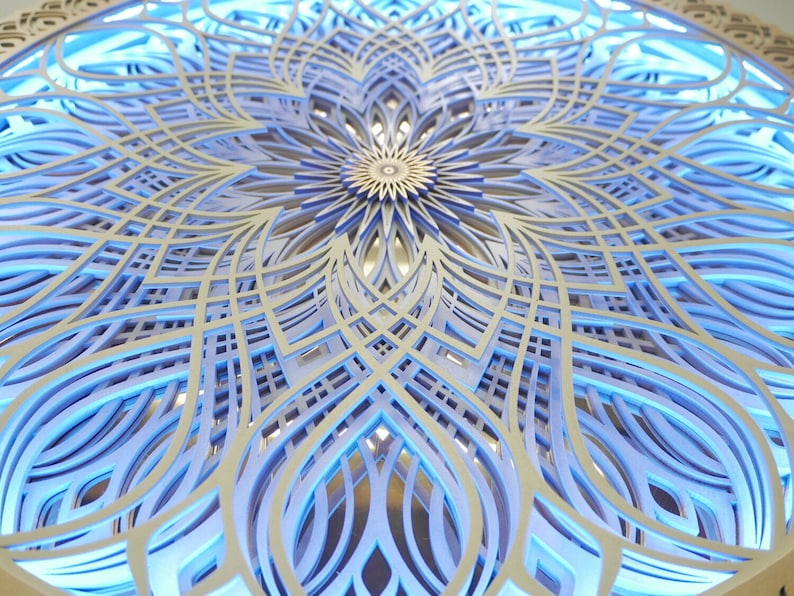 May include: A circular wall art piece featuring intricate, multi-layered laser-cut wood panels arranged in a symmetrical mandala pattern. The design is illuminated from behind with a bright blue light, highlighting the depth and geometric cutouts.