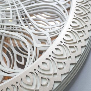 May include: A circular wall art piece featuring multiple layered, laser-cut white panels with intricate, symmetrical, curved geometric patterns. The design creates a sense of depth through stacked cutouts, revealing subtle wood-toned layers beneath.