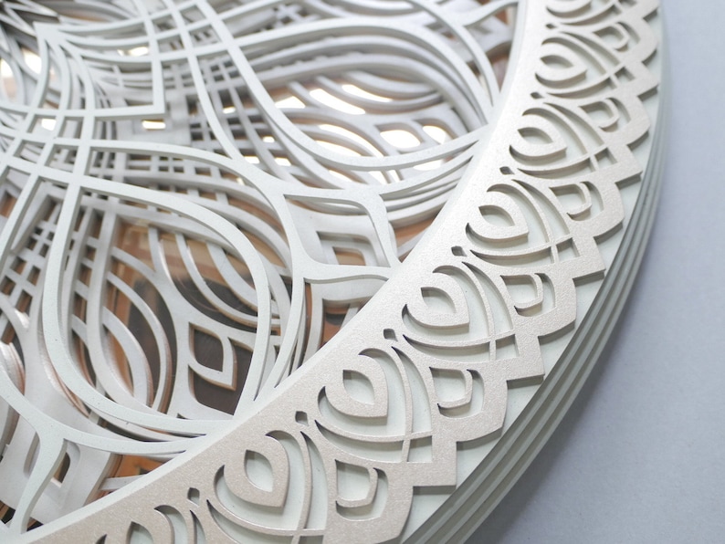 May include: A circular wall art piece featuring multiple layered, laser-cut white panels with intricate, symmetrical, curved geometric patterns. The design creates a sense of depth through stacked cutouts, revealing subtle wood-toned layers beneath.