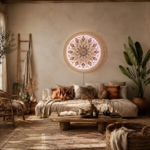 May include: A circular wall-mounted light fixture featuring an intricate, laser-cut wooden mandala design with a warm, glowing backlight. The piece is mounted on a textured beige wall above a low-profile sofa, serving as a decorative focal point.