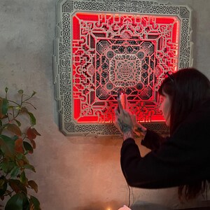 Pu&ograve; includere: Un'opera d'arte da parete quadrata in legno a strati con intricati motivi geometrici e retroilluminazione a LED rossa. L'opera &egrave; montata su una parete grigia, creando un contrasto sorprendente. Il design presenta una complessa disposizione simmetrica.
