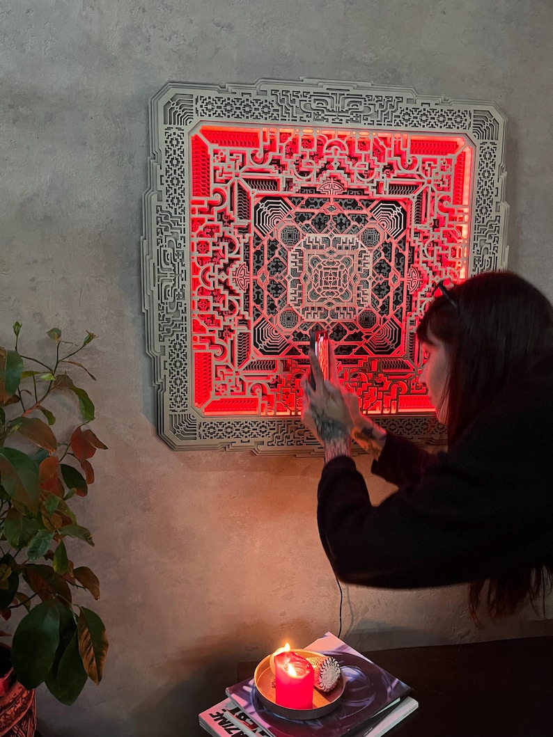 Pu&ograve; includere: Un'opera d'arte da parete quadrata in legno a strati con intricati motivi geometrici e retroilluminazione a LED rossa. L'opera &egrave; montata su una parete grigia, creando un contrasto sorprendente. Il design presenta una complessa disposizione simmetrica.