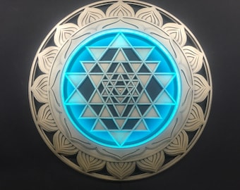 May include: A decorative wall art piece featuring a Sri Yantra design. The artwork has a layered, metallic appearance with a central geometric pattern illuminated by a bright blue light. The outer ring has a lotus flower design.