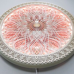 May include: A circular wall-mounted light fixture featuring intricate, laser-cut layered wood panels in a cream color. The design displays a symmetrical mandala pattern with a soft, warm pink glow emanating from behind the cutouts.