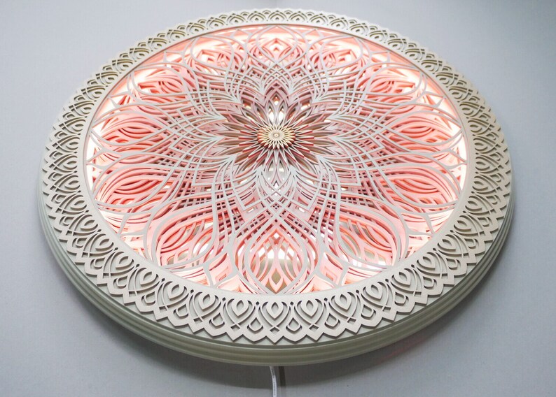 May include: A circular wall-mounted light fixture featuring intricate, laser-cut layered wood panels in a cream color. The design displays a symmetrical mandala pattern with a soft, warm pink glow emanating from behind the cutouts.