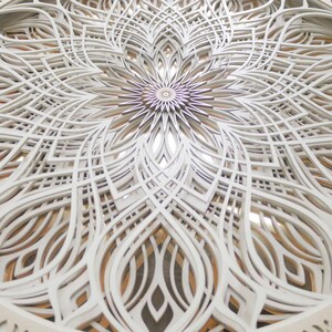 May include: A circular wall art piece featuring multiple layers of white laser-cut wood arranged in a complex, symmetrical mandala pattern. The intricate design consists of repeating geometric shapes and curved lines, revealing warm wood tones.