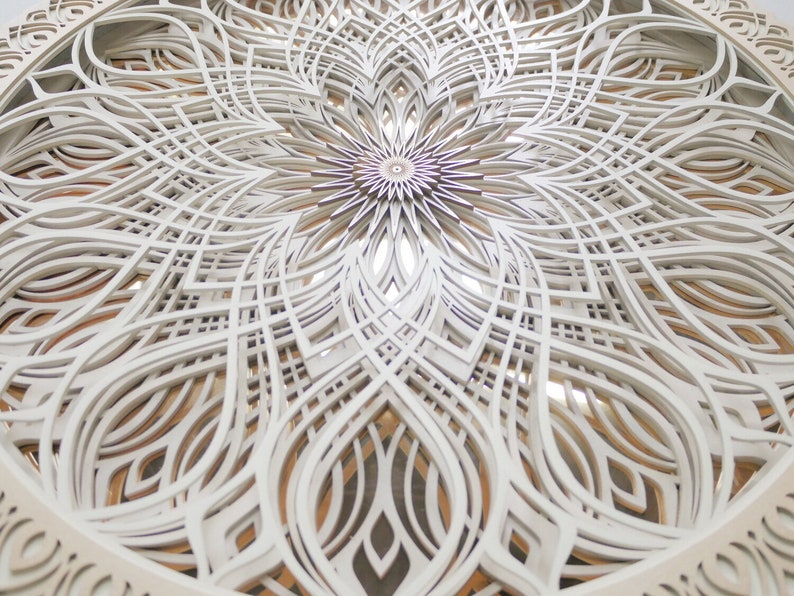 May include: A circular wall art piece featuring multiple layers of white laser-cut wood arranged in a complex, symmetrical mandala pattern. The intricate design consists of repeating geometric shapes and curved lines, revealing warm wood tones.