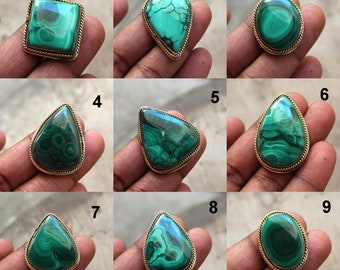 May include: Nine malachite gemstone rings with gold-colored metal settings. The malachite stones vary in shape, including square, oval, and teardrop. The green stones have unique swirling patterns. The rings are displayed on a hand.