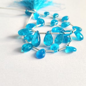May include: A collection of bright blue gemstone beads in teardrop and oval shapes, strung on silver wire. A matching blue tassel is visible in the background. The translucent beads have internal patterns. The beads are measured in centimetres.
