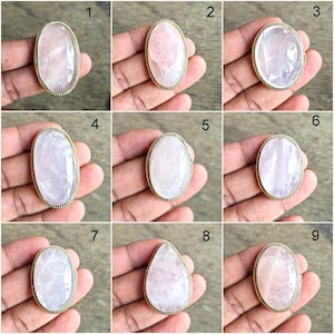 Puede incluir: Colección de nueve anillos de cuarzo rosa con engastes de metal dorado. Las piedras preciosas varían en forma, incluyendo ovaladas y en forma de lágrima, y muestran tonos de rosa y blanco. Los anillos se muestran en una mano.