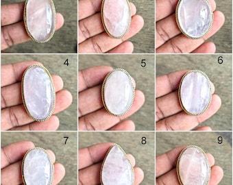 May include: A collection of nine rose quartz rings with gold-colored metal settings. The gemstones vary in shape, including oval and teardrop, and display shades of pink and white. The rings are displayed on a hand.