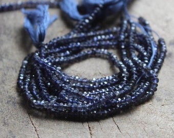 May include: A strand of dark blue faceted gemstone beads. The beads are small and round, and they are strung on a thin thread. The beads are arranged in a loose coil, and they are lying on a wooden surface.